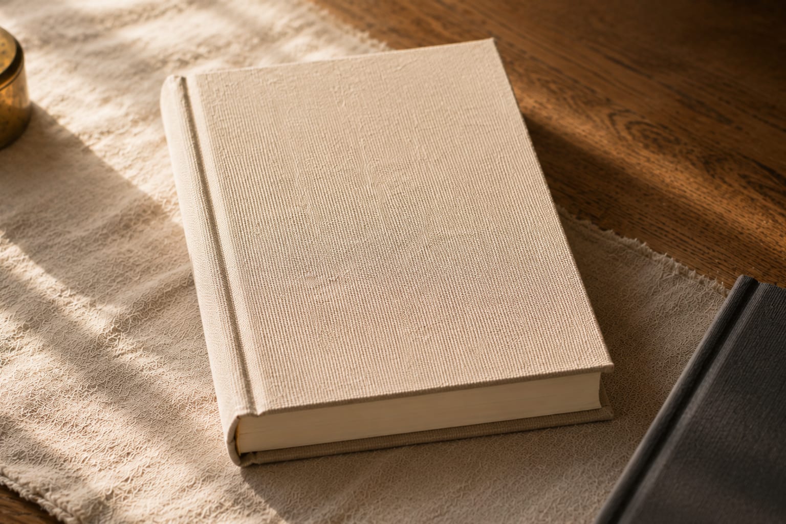 A close three-quarter detail of a casewrap hardcover resting on raw linen, the printed cloth wrapping the spine and boards visible at the spine fold, warm afternoon window light catching the woven texture of the cloth, a second closed book at the lower edge of frame in soft defocus.